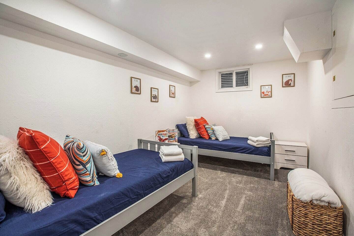 Habitación tipo dormitorio con dos camas individuales, cada una con colchón azul y varias almohadas. Hay cuadros enmarcados en la pared, una ventana pequeña y un equipo de aire acondicionado blanco.