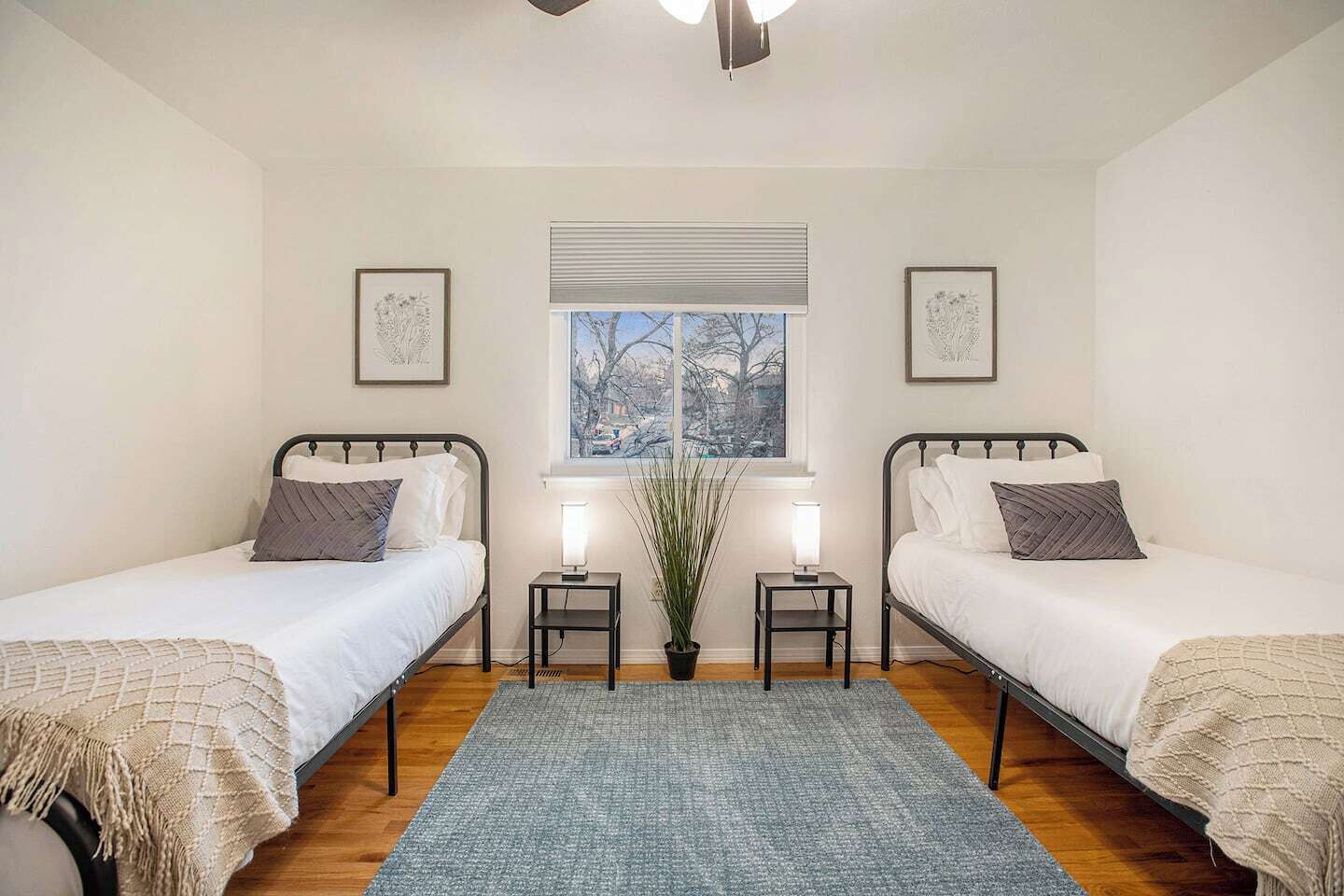 A bedroom with two single beds, a window with blinds, a potted plant, and bedside lamps.