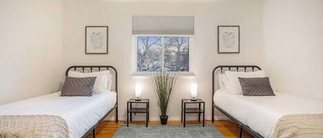 A bedroom with two single beds, a window with blinds, a potted plant, and bedside lamps.