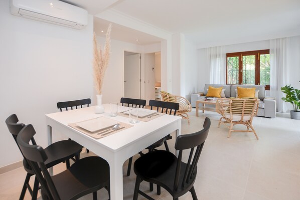 Un salon moderne avec une table à manger blanche, dressée pour quatre personnes, entourée de chaises noires. On aperçoit un canapé avec des coussins jaunes et un fauteuil en osier au fond de la pièce.
