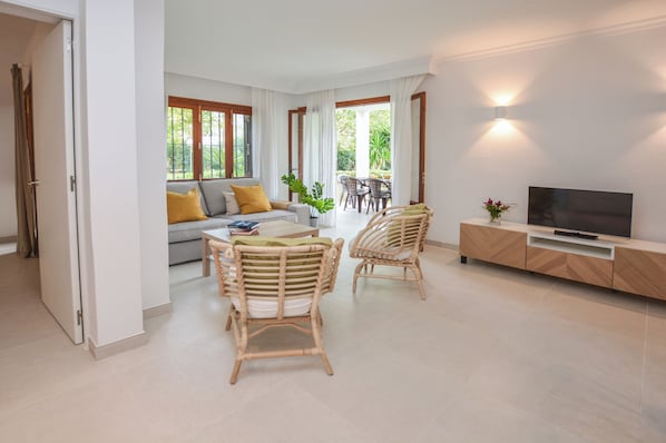 Een moderne woonkamer met een bank, twee rieten stoelen, een eettafel en een televisie op een houten dressoir.