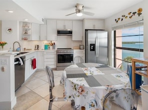 A modern kitchen with white cabinetry, a stainless steel refrigerator, a microwave, and a stove. There is a dining table with a floral tablecloth and chairs. A ceiling fan is visible, and the room has a view of the ocean through large windows.