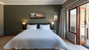 Ein großes Bett mit weißen Leinen in einem Hotelzimmer, ein Nachttisch mit Lampe, ein Gemälde an der Wand und eine Glastür zum Außenbereich.
