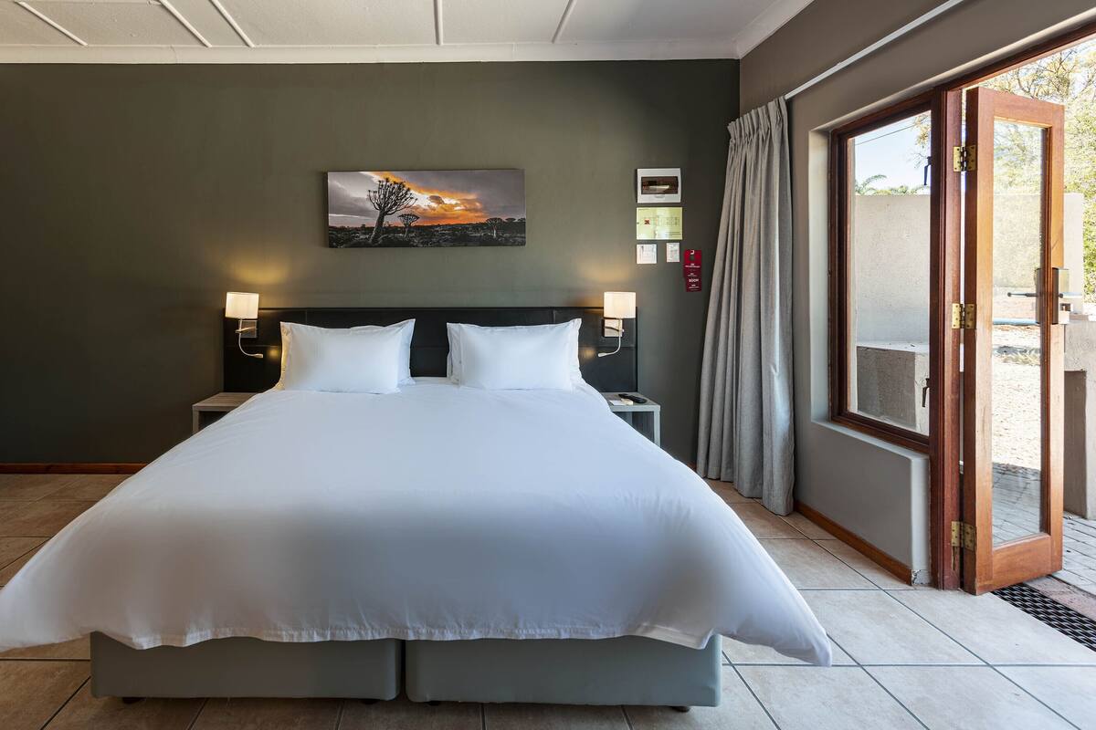 A large bed with white linens in a hotel room, a nightstand with a lamp, a painting on the wall, and a sliding glass door to the outside.