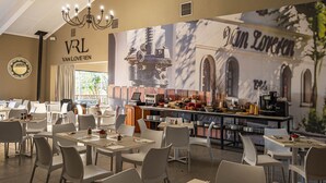 Ein Restaurant mit mehreren Tischen und Stühlen, einer Bar mit verschiedenen Speisen und einer Kaffeemaschine, sowie einem Wandgemälde.