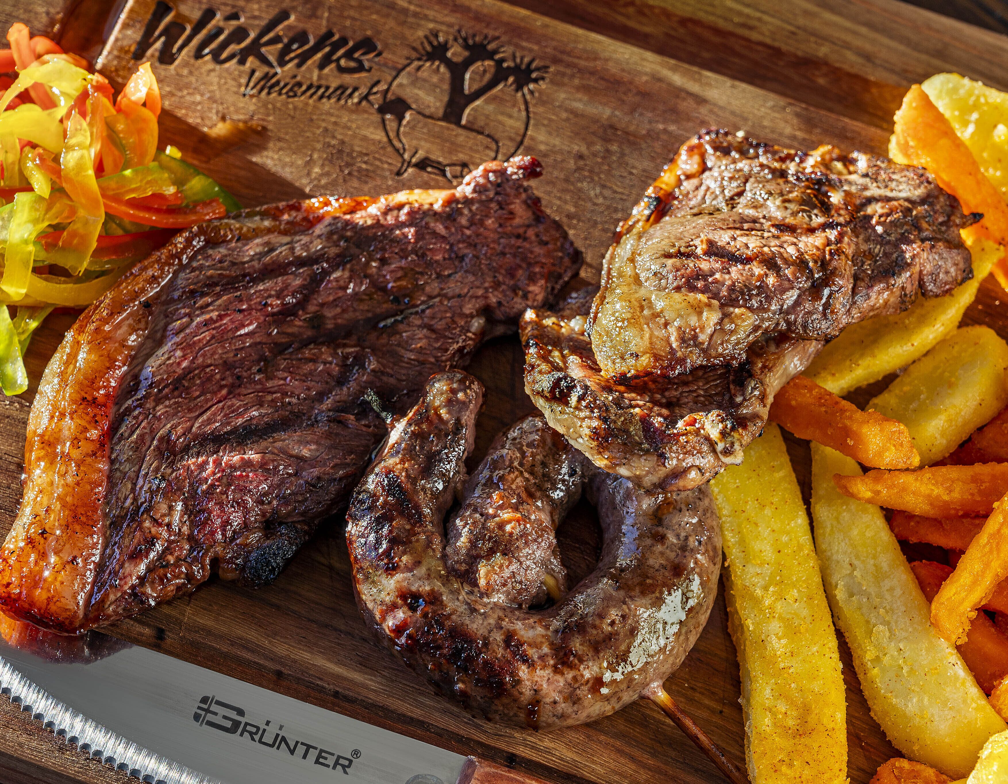 A wooden cutting board with grilled steak, sausage, and French fries.