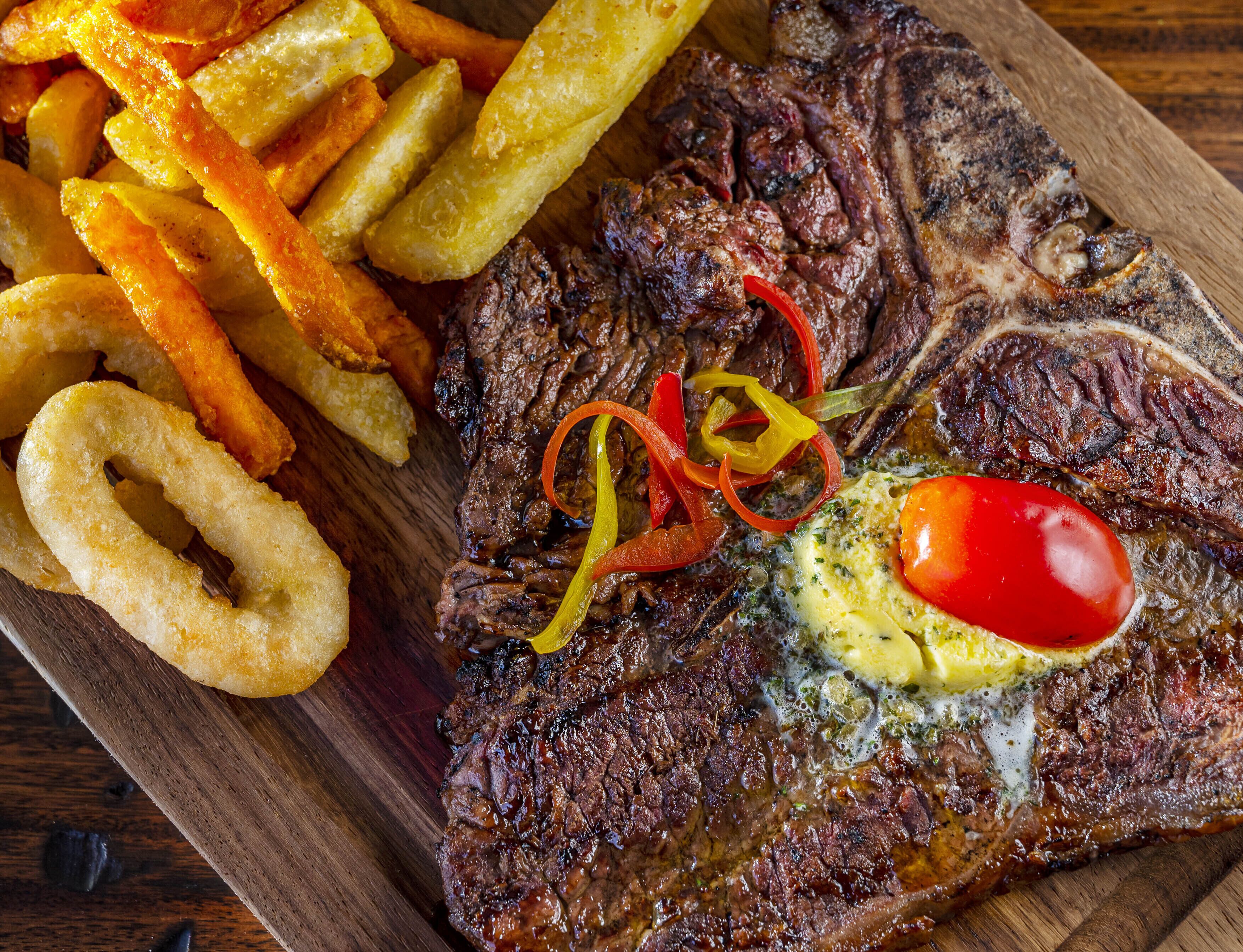 Un bife acompañado de aros de cebolla, papas fritas y una guarnición de tomate cherry y pimiento, todo sobre una tabla de madera.