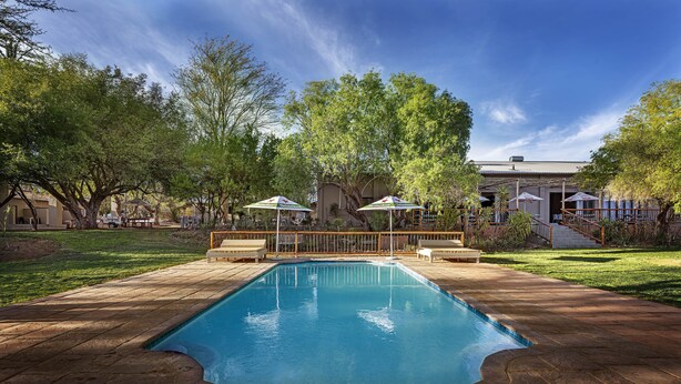 Ein Swimmingpool mit Holzterrasse, umgeben von Bäumen und Sitzgelegenheiten mit Sonnenschirmen.