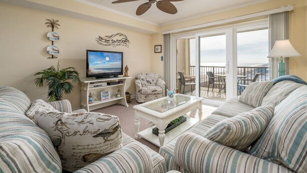A living room with striped sofas, a ceiling fan, a television, and a balcony with a view.