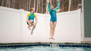 Two people jumping into a swimming pool with a white fence and trees in the background.