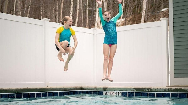 Two individuals jumping into a swimming pool from a white fence.