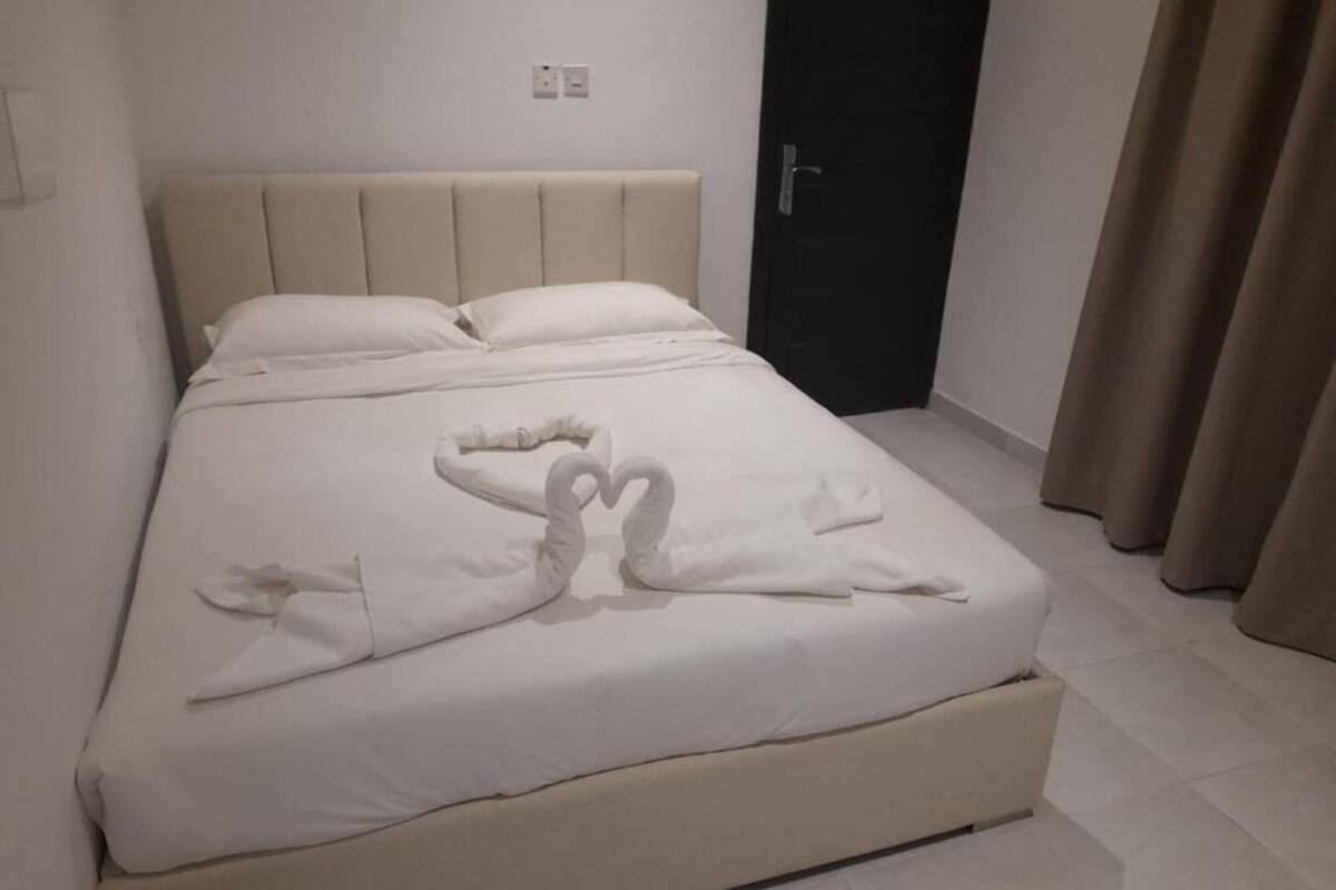 A neatly made bed with a heart-shaped towel arrangement, a dark door, and beige curtains.