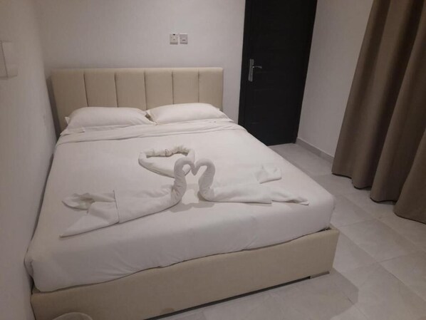Una cama bien hecha con un arreglo de toallas en forma de corazón, una puerta oscura y cortinas color beige.