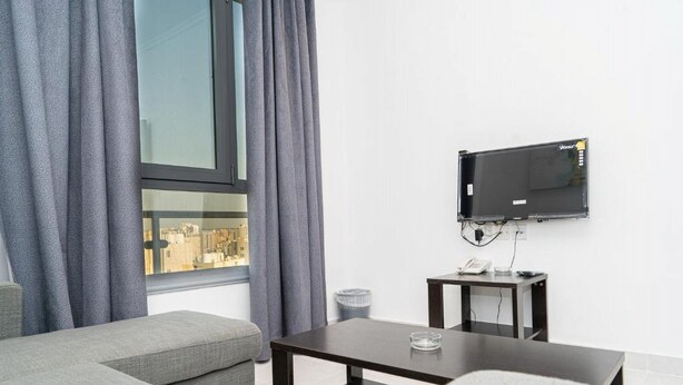 A modern hotel room with a large window, grey curtains, a flat-screen TV mounted on the wall, a small table with a glass, and a sofa. - Atlantis Hotel Suites (Salmiya)