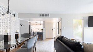 Ein modernes Wohnzimmer mit einem Glastisch, grauen Stühlen, einem schwarzen Ledersofa und einer Küche im Hintergrund.