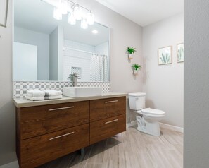 A modern bathroom with a wooden vanity, white sink, and toilet. There are two potted plants on the wall and framed artwork above the toilet. - Modern Chic Condo In Midtown w/ Pool & Hot Tub (Phoenix)