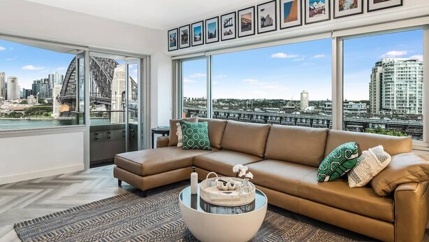 Una sala de estar moderna con un ventanal grande, un sofá de cuero marrón y vistas a un horizonte urbano que incluye un puente.