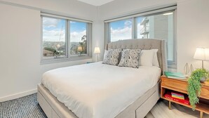 Un dormitorio moderno con una cama grande, dos ventanas, una mesita de noche con lámpara y una estantería.