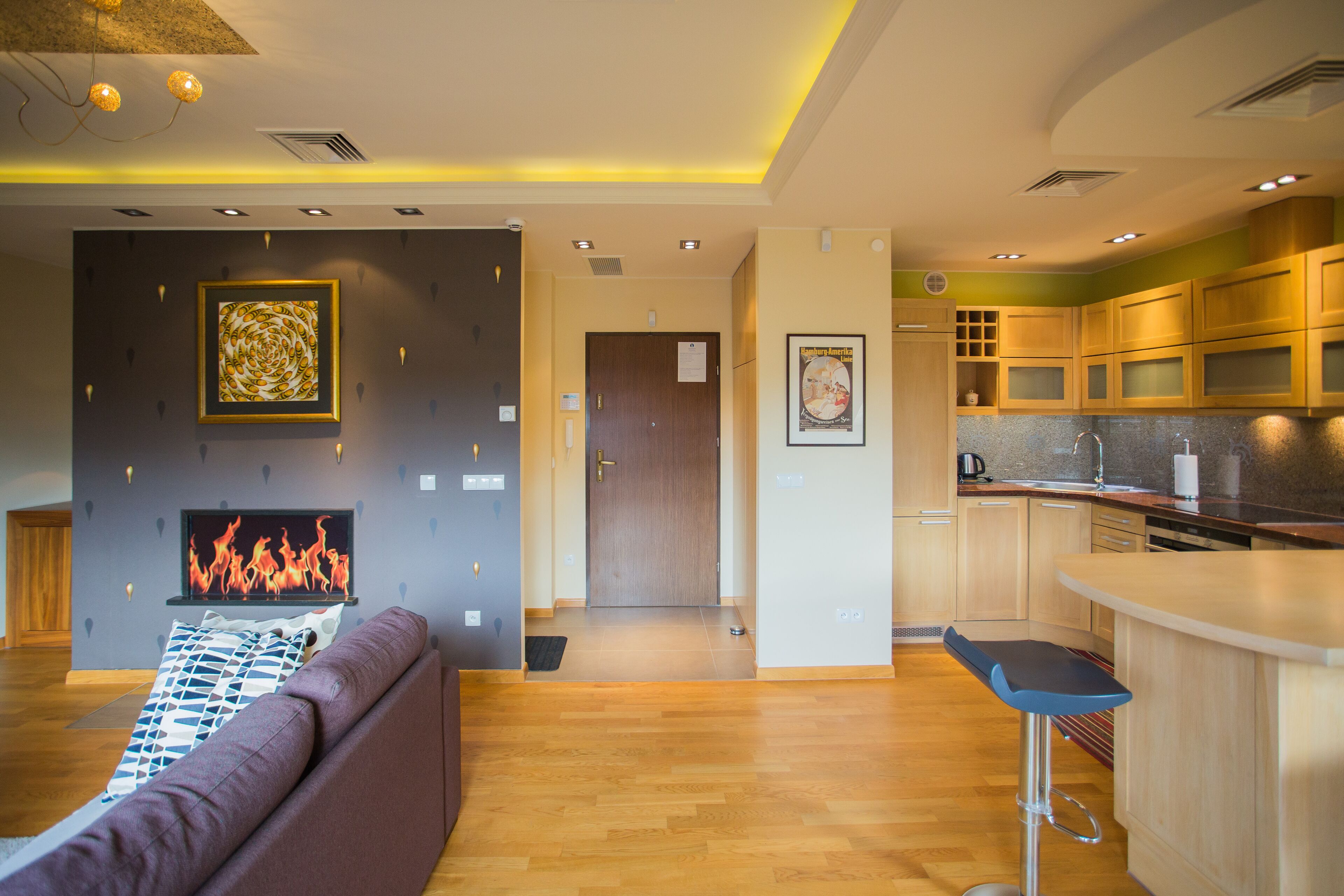 A modern living room with a fireplace, a kitchen area with wooden cabinets, and a bar stool.