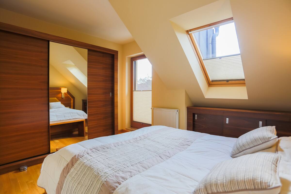 A bedroom with a skylight, a bed with white bedding, a wooden wardrobe, and a mirror reflecting the room.
