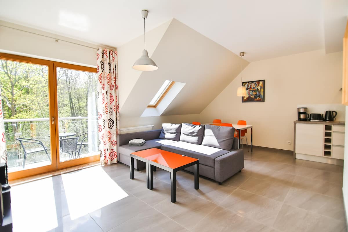 A modern living room with a sofa, a coffee table, and a dining area with a table and chairs. There is a balcony with a view of trees.