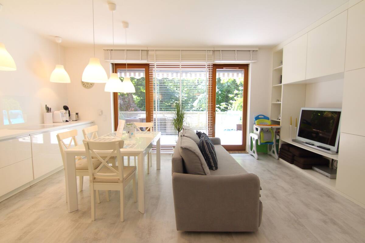 A modern kitchen with white cabinets, a dining table with chairs, a sofa, and a television.