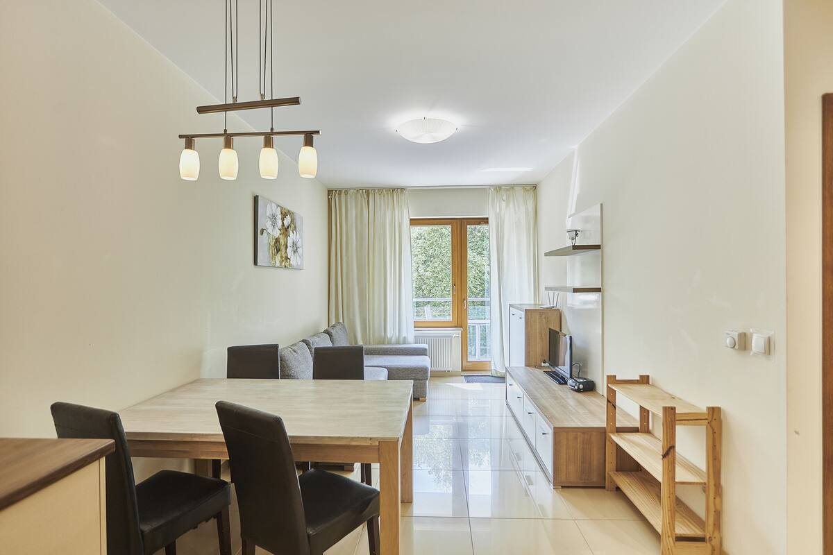 A modern kitchen with a dining area, wooden furniture, and a view of greenery outside.