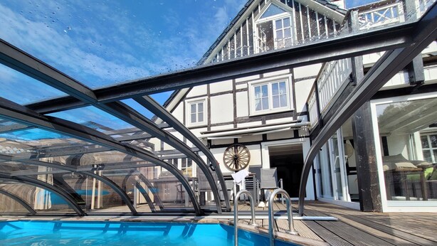 Una piscina con cerramiento de vidrio, una terraza de madera y un edificio con balcón al fondo.