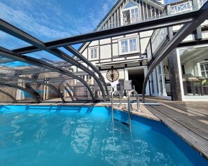 Una piscina con cerramiento de vidrio, una terraza de madera y un edificio con balcón al fondo.