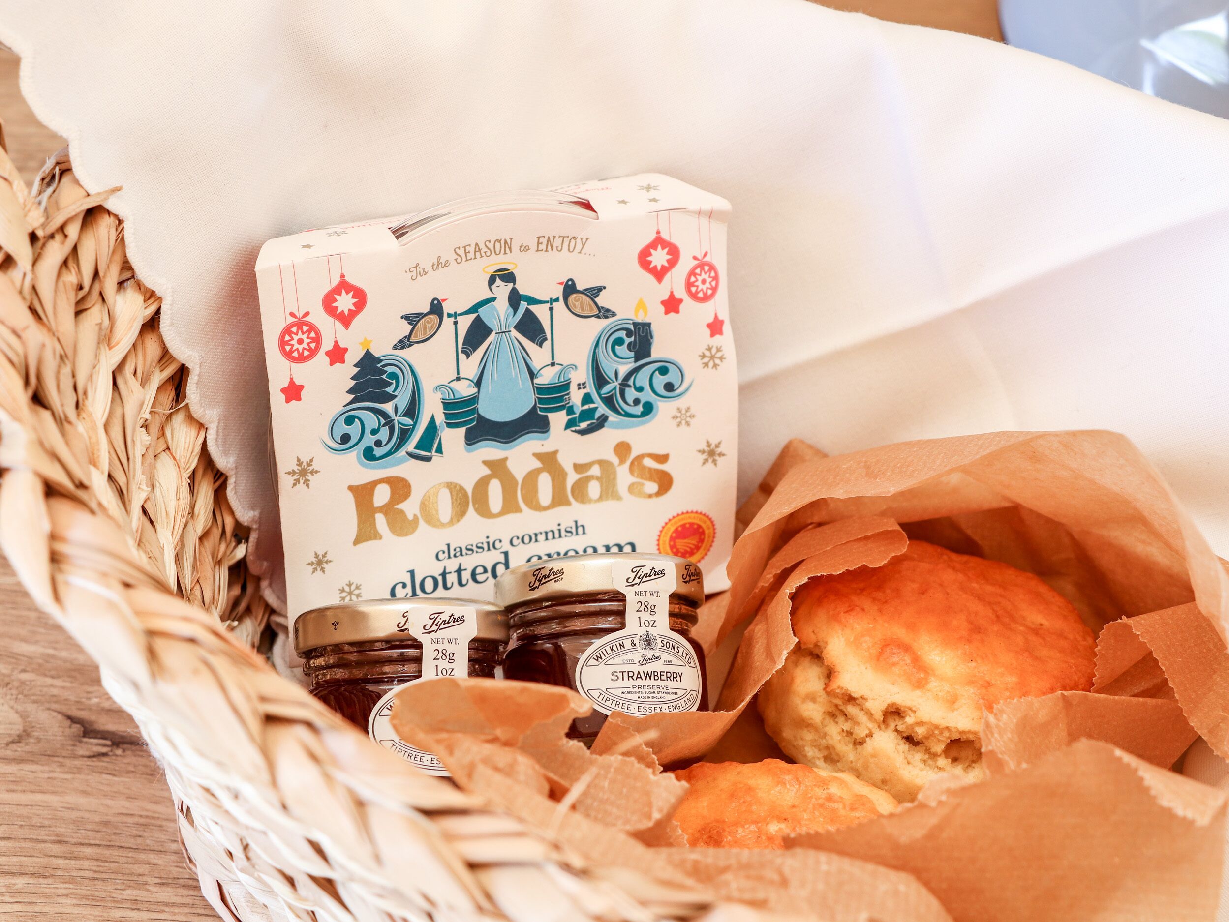 A basket containing a loaf of bread, a jar of Rodd's classic cornish clotted cream, and a box of Tiptree brand jam.