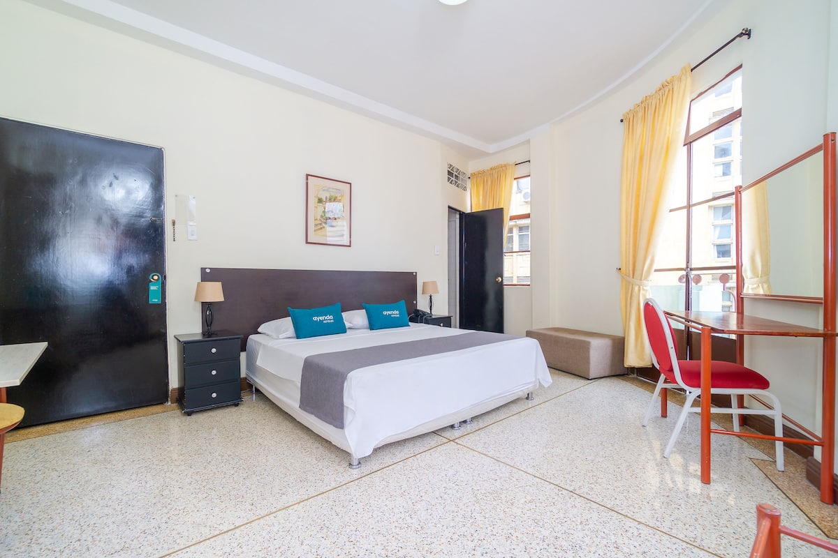 Quarto de hotel com cama, escrivaninha, cadeira, guarda-roupa e janela com cortinas.