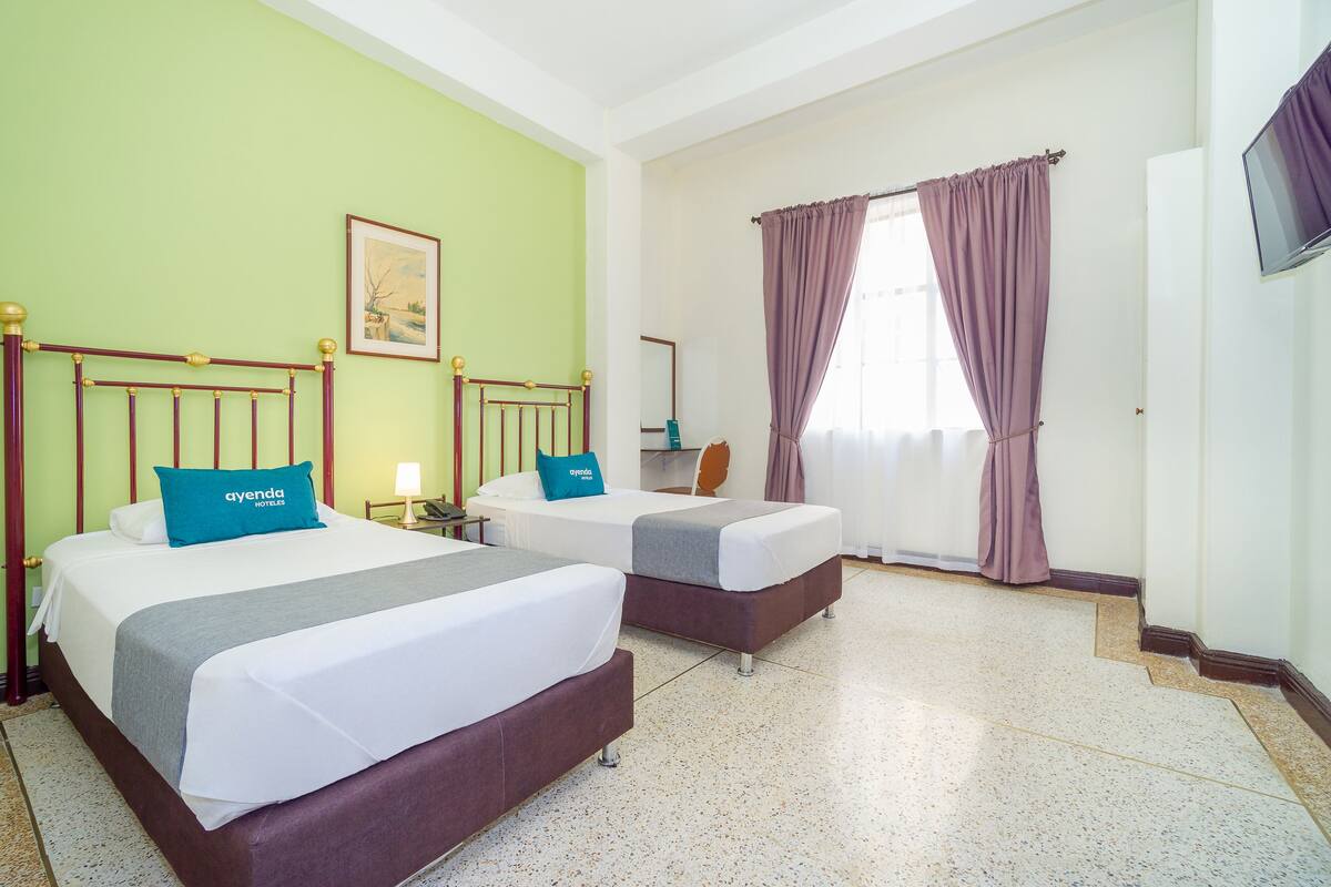 Quarto de hotel com duas camas, janela com cortinas roxas, televisão na parede e um quadro na parede.