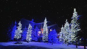 Schneebedeckte Landschaft mit beleuchteten Weihnachtsbäumen und einem Gebäude im Hintergrund.