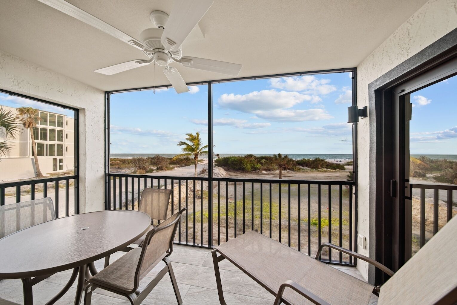 A balcony with a table and chairs, a ceiling fan, and a view of the beach and ocean.
