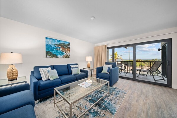 A modern living room with a blue sofa, a glass coffee table, and a balcony with outdoor furniture. - Pelicans Roost 107: Corner Unit with Gulf Views! (Sanibel)