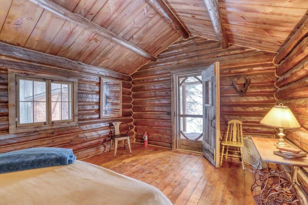 A log cabin room with a bed, two chairs, a table with a lamp, and a large window.