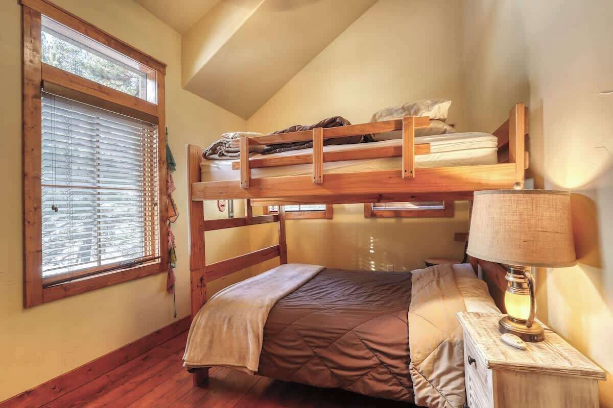 Quarto estilo dormitório com beliches, janela com persianas, luminária de cabeceira e piso de madeira.