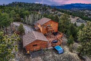 A two-story log cabin with a deck and hot tub, surrounded by forested hills. - Newly Furnished Mountain Home - Private Hot Tub- Fooseball - Unbeatable Views (Bailey)