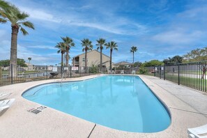 A rectangular pool with a tiled edge, surrounded by a black metal fence, palm trees, and a building with a flat roof in the background. - Dock Holiday 2 Bedroom Condo by Redawning (Rockport)