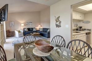 A modern living room with a glass dining table, blue sofas, and a kitchen area with white cabinets and a clock. - Dock Holiday 2 Bedroom Condo by Redawning (Rockport)
