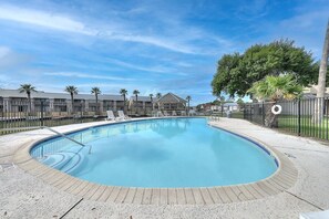 A swimming pool with lounge chairs, surrounded by a fence, palm trees, and a building in the background.