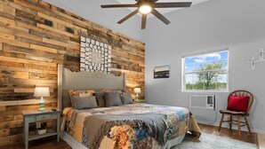 A bedroom with a wooden wall, a large bed with a quilt, a nightstand with a lamp, a window with a view, and a ceiling fan. - Fairway Heights 2 Bedroom Condo by RedAwning (Branson)
