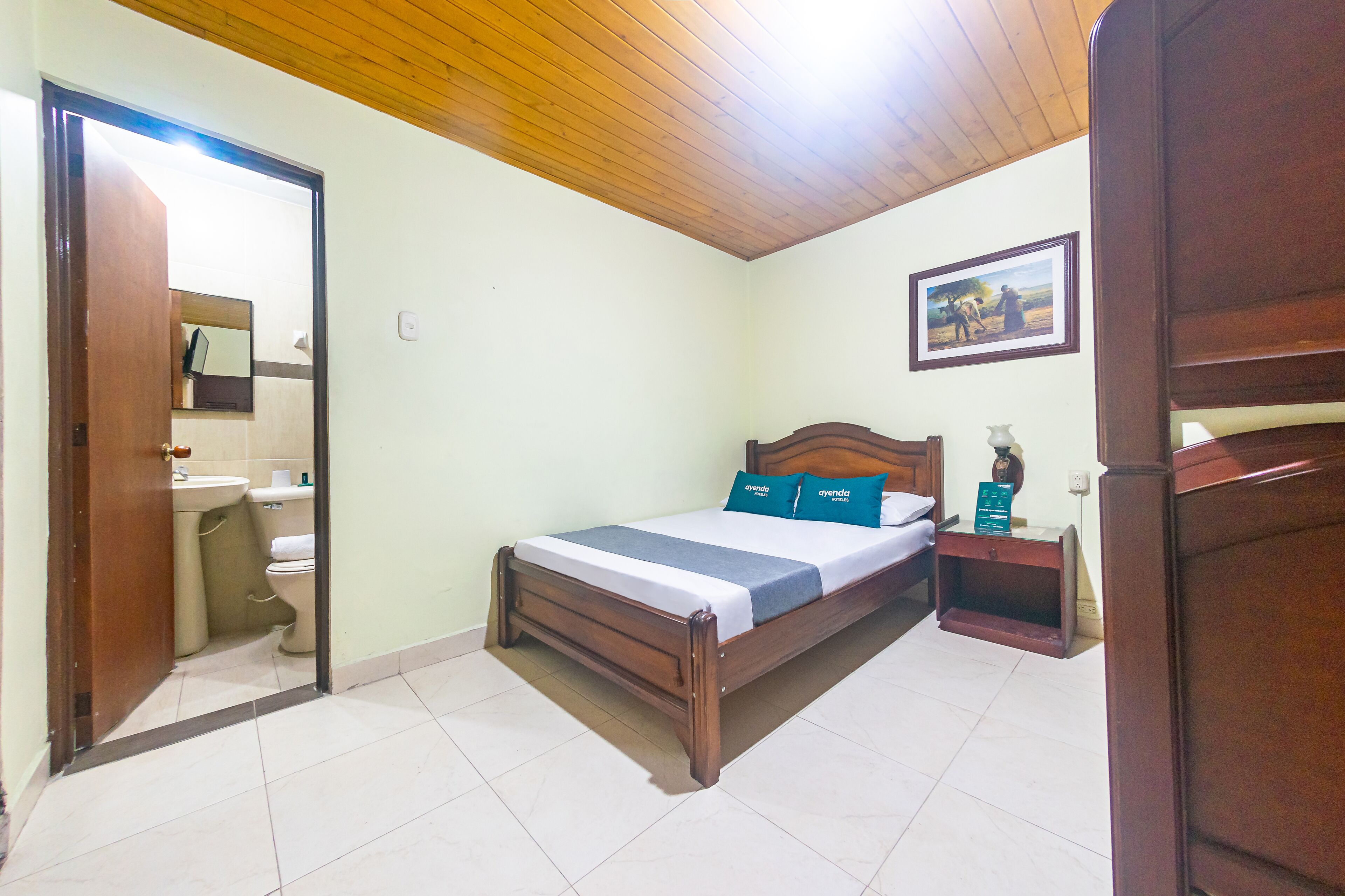 Un dormitorio con una cama de madera, una mesita de noche, una pequeña mesa y un baño visible a través de una puerta abierta.