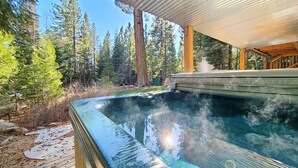 Een hottub met een houten terras en een pergola erboven, omringd door bomen.