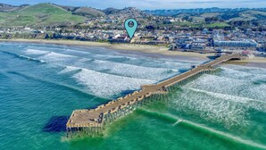 An aerial view of a coastal town with a wooden pier extending into the ocean, surrounded by residential buildings and hills in the background.