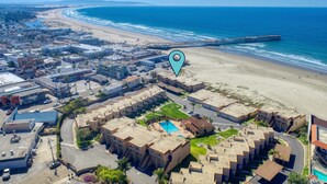 An aerial view of a coastal town with a beachfront hotel, featuring a pool, a pier, and a clear view of the ocean.