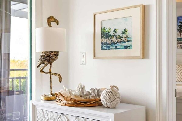 A white console table with a framed painting of a tropical beach scene, a decorative bird lamp, and a woven basket with natural decor.