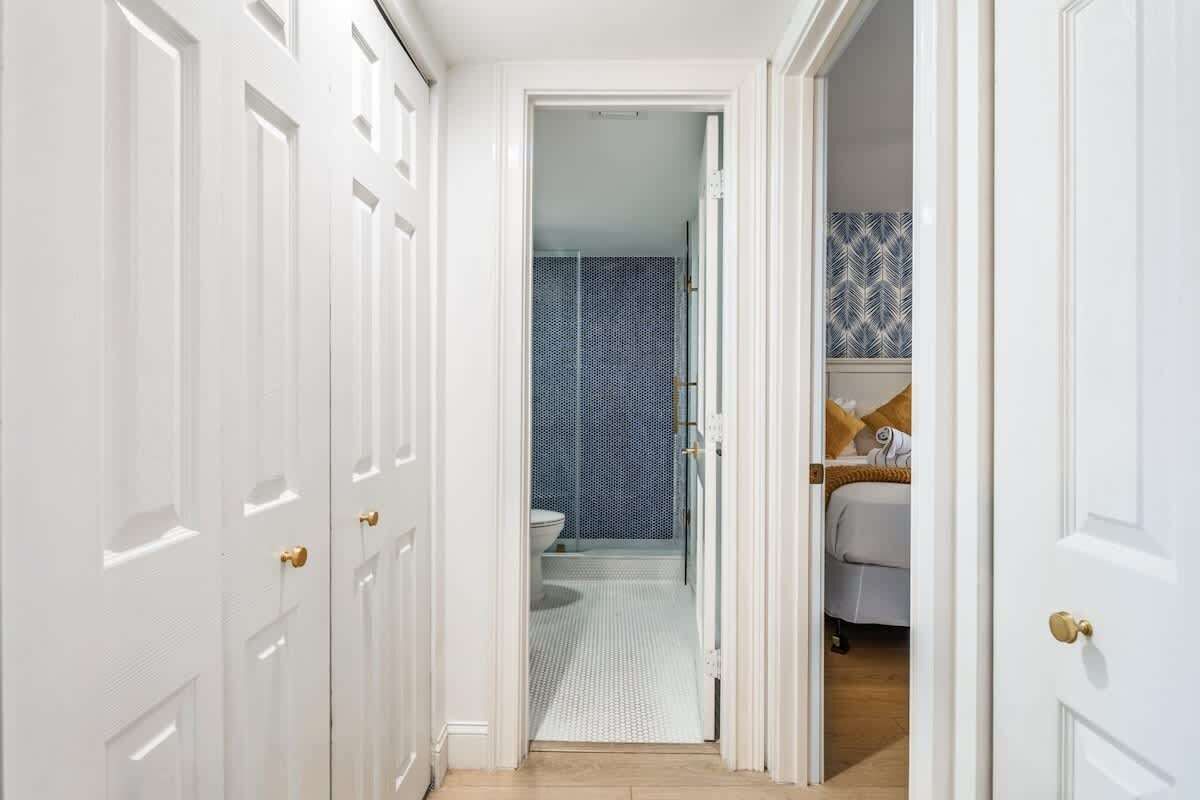 A hallway with white doors leading to a bathroom with a patterned shower wall and a bed with a patterned bedspread.