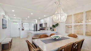 A modern kitchen with white cabinets, a central island, and a dining area with wicker chairs and a chandelier. - Island Time | Luxury Beach Condo w/ Pool & Hot Tub (Key West)