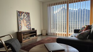 A living room with a laptop on a round table, a sofa, a chair, a bookshelf, and a large window with a mountain view. - Apt With Panoramic View Near Vauban City (Briançon)